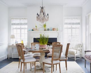 All white, bright dining room, black flooring, white painted fireplace with decorative ornaments on mantel, wooden dining table with cream painted column, matching wooden dining chairs with woven backs and upholstered seats, striped cream rug below table, wooden bench and seat with upholstered seats and cushions on edge of the room
