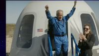 an older man in a blue flight suit emerges from a white space capsule with arms raised in triumph