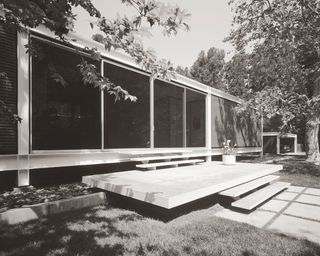 Black and white image of Rosen House, Los Angeles, grass lawn, large stone slab steps leading up the house, large floor to ceiling glass panels, trees to the left and right, stone paving under the steps, clear sky