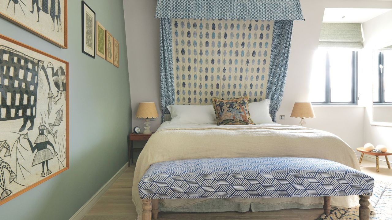 A blue bedroom with a patterned bed canopy and ottoman, framed art on the wall, matching beige scalloped lamps either side of the bed with white pillows, beige blanket and patterned cushion, and a bright window with open sage green blind with a small wooden table beneath