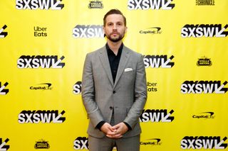 Logan Marshall-Green standing in front of a bright yellow backing board wearing a grey suit