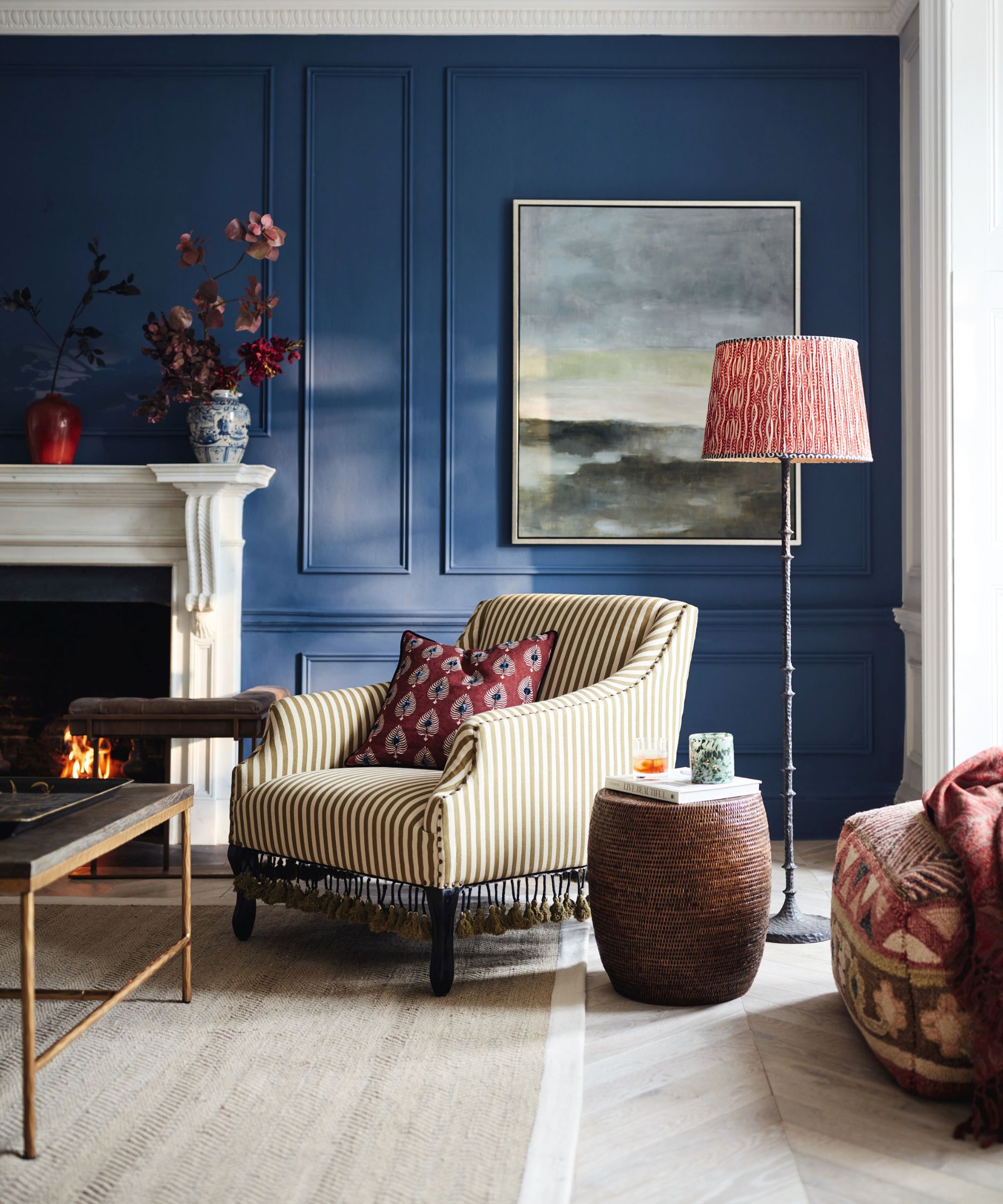 A deep navy panelled living room with a lit white marble fireplace, gold-striped tasselled armchair, red patterned floor lamp, wicker side table and a muted abstract landscape painting above.