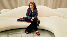 A woman dressed in a casual chic leather jacket with red embroidery and red leather mules sits on a sculptural sofa.