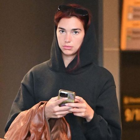 Dua Lipa wearing a black hoodie, brown leather jacket, and black sweatpants at JFK airport.