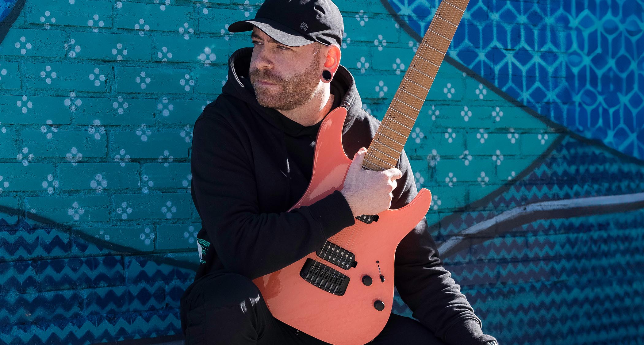 Aaron Marshall of Intervals holds a peach-coloured S-style as he is photographed against a green and blue patterned wall.