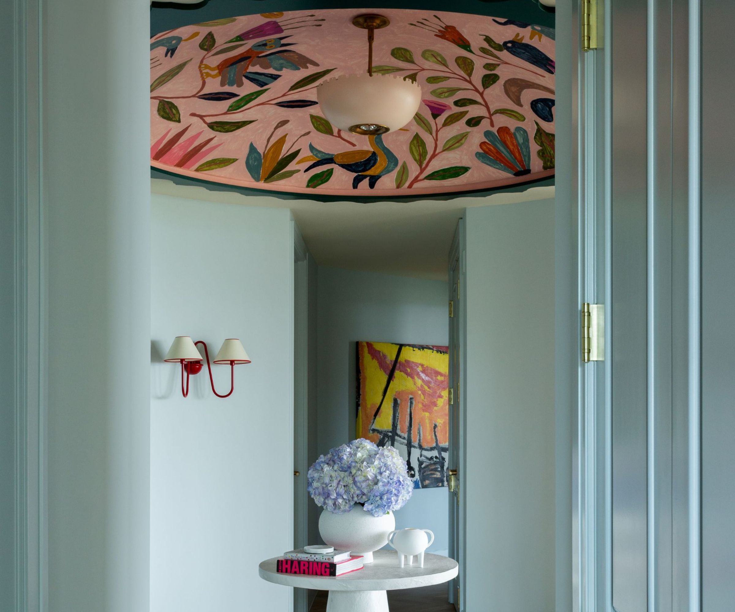 Pale blue entryway with dark teal double doors, two red sconces on walls, a large pink floral ceiling mural, a round, small white table in the centre with a vase and books, and parquet wooden floors