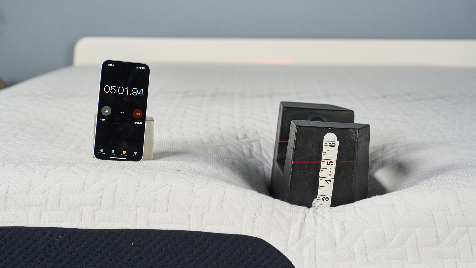 A closeup of the Nectar Premier Memory Foam Mattress on a bed frame in our sleep studio during the edge support test. A 25kg weight is balanced on the edge of the bed with a tape measure attached to the side. A red laser line is directed at the tape, indicating how far the weight has sank into the mattress. To the left, a phone displaying a stop watch is balanced. It indicates the test has been going on for 5 minutes 1 second.