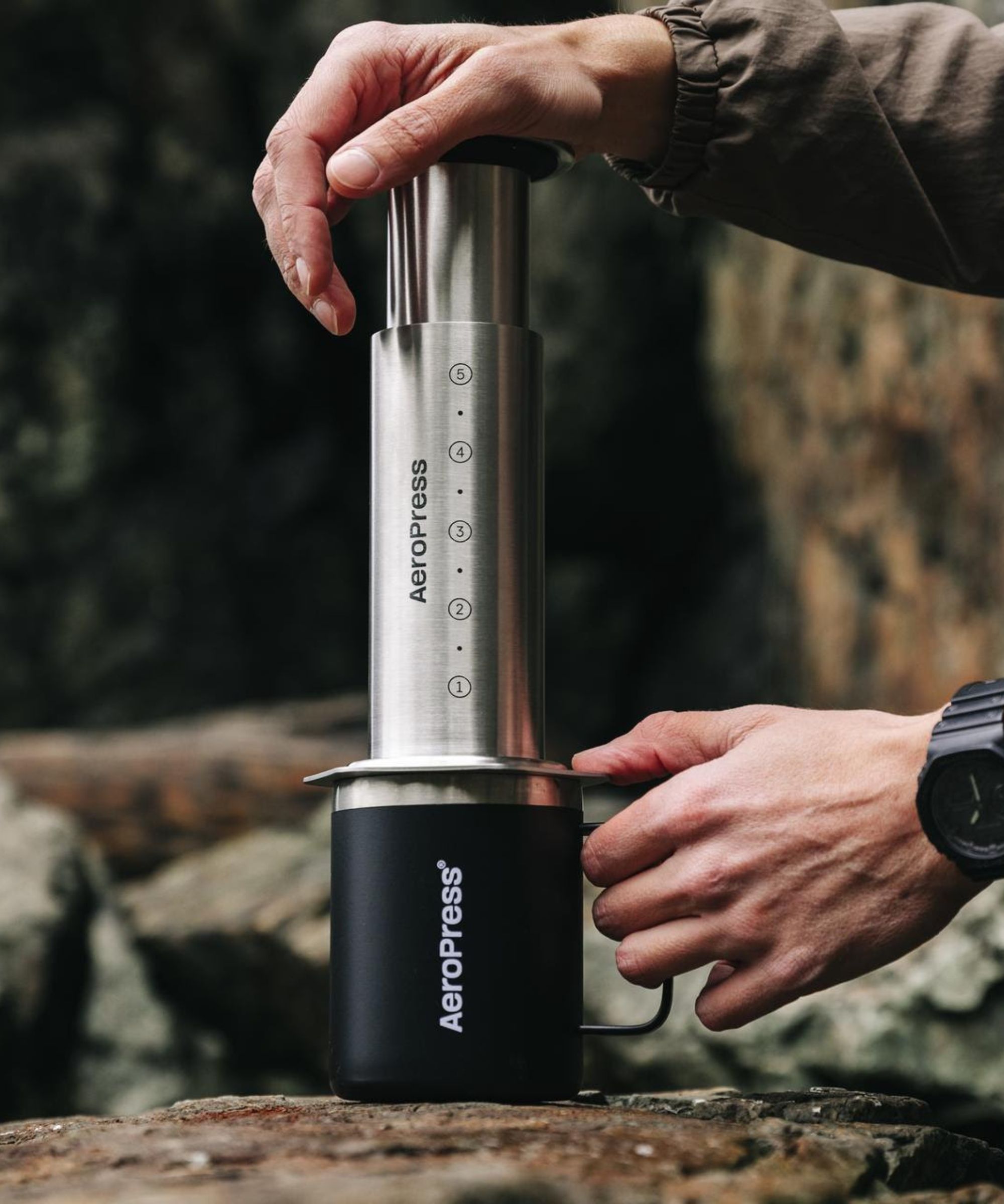 The stainless steel AeroPress being used by a Caucasian man to extract coffee into a black branded travel mug on a wooden log.