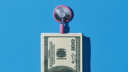 A pink stethoscope and a pile of hundred-dollar bills against a blue background.