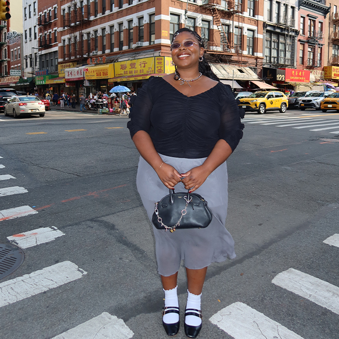 Chichi is wearing a black top, gray skirt, Mary Janes, white socks, and holding a black handbag while posing in the streets of NYC. 