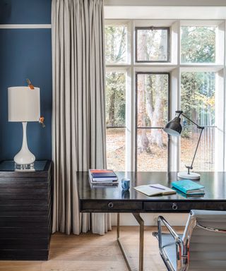 Study with a modern wood desk and pale grey office chair and a large window, pale grey carpet and curtains and blue walls