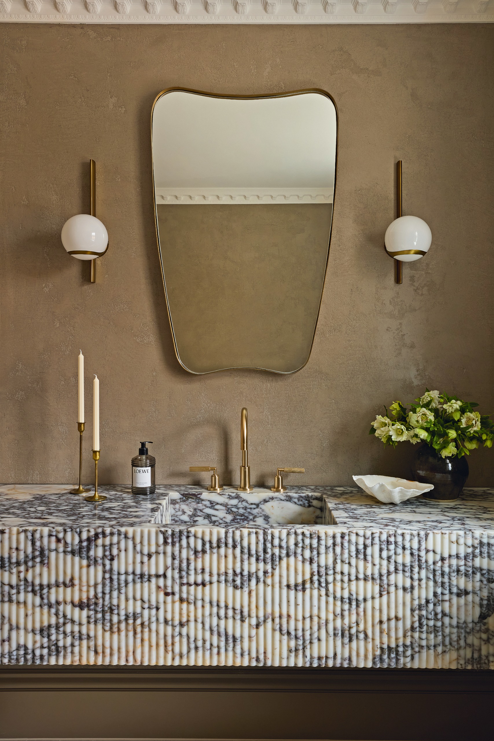Powder room with taupe tadelakt walls, cream and grey ribbed marble sink, brass taps, brass and opal glass wall lights and curved mirror