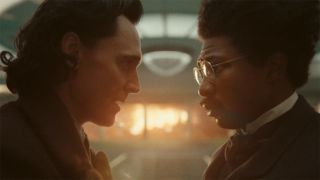 Image from the Marvel T.V. show Loki, season 2 episode 6. Close up of two men facing each other having a serious discussion. The man on the left has dark slicked back hair and is wearing a jacket. The man on the right has thick dark hair, round gold glasses and is wearing a suit and cravat. Sunlight glows from the window behind them.