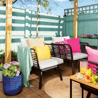 Garden decking area with blue painted fence, black outdoor furniture, pink and yellow cushions and round natural woven rug