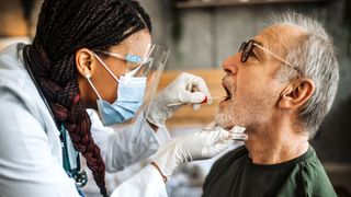 Profile view of a doctor peering into the open mouth of the patient while holding the patient's chin and placing a long cotton swab into the patient's mouth. 