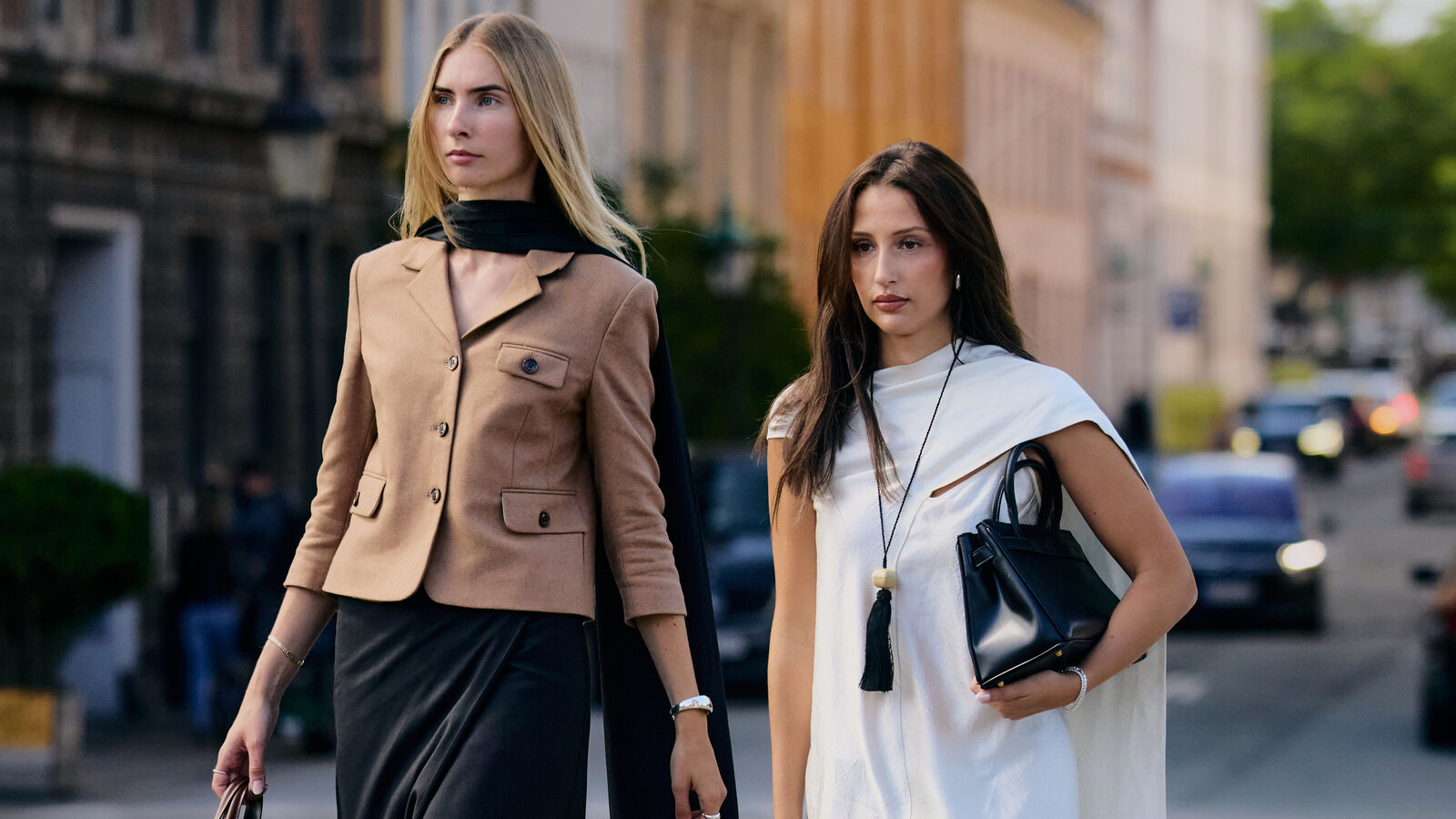 Two women are walking and posing. One wears brown jacket and black pants. The other wears a white dress with heels and black handbag. 