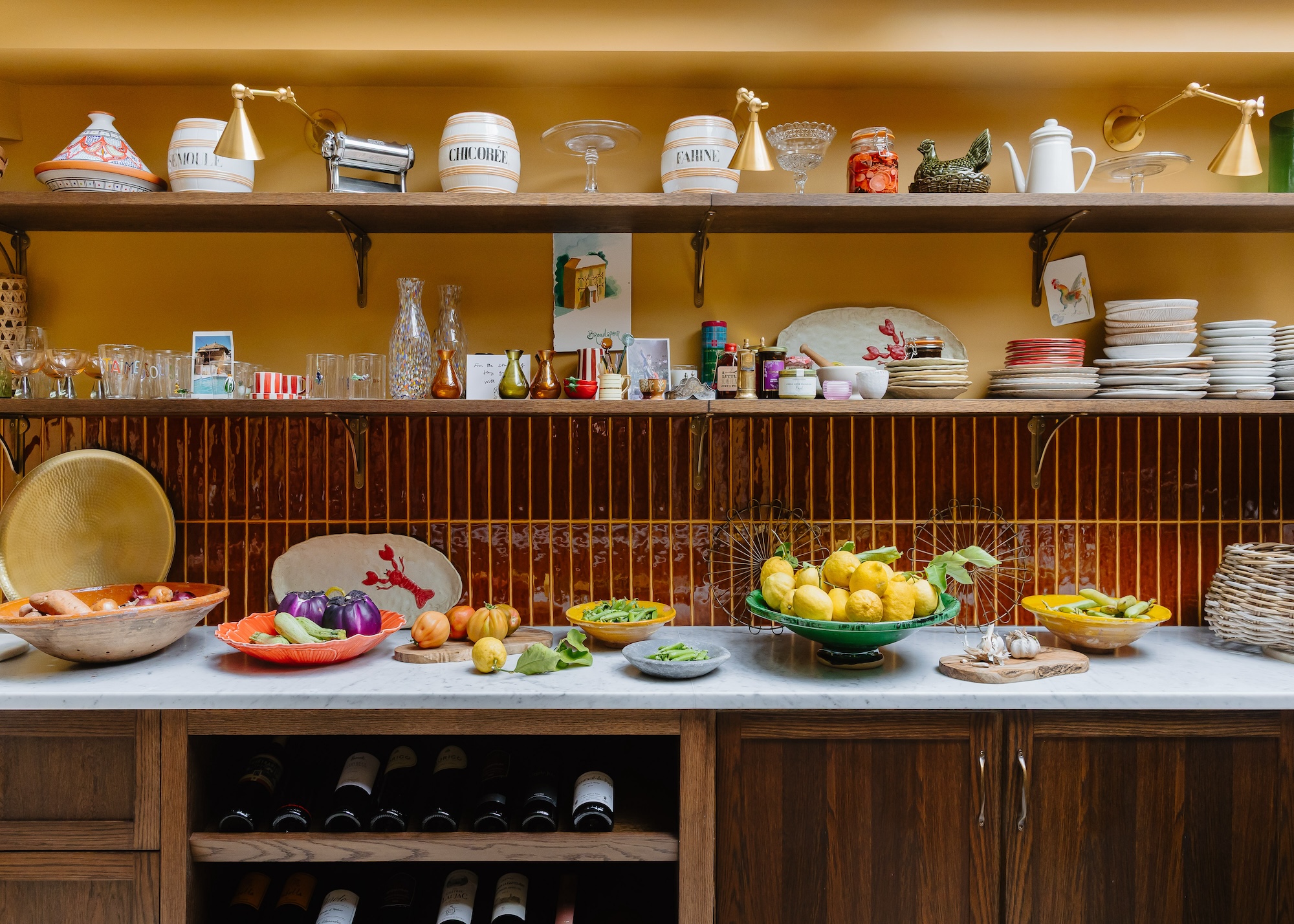Open shelves in a chef's kitchen with dishes, plates as well as other kitchen essentials. Beneath the shelf is a countertop with lots of fruits and vegetable bowls. There is also a view of the wine storage compartment beneath the counter