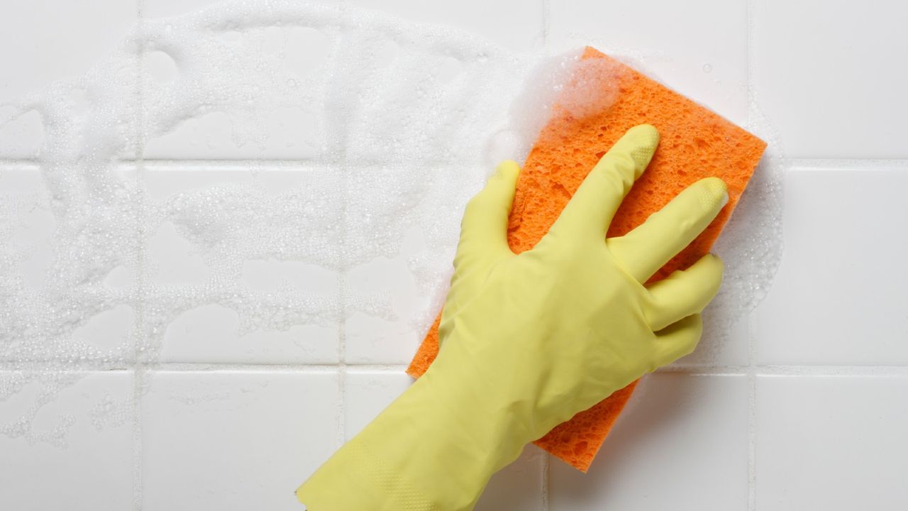 Person wearing a yellow rubber glove wiping down a bathroom wall