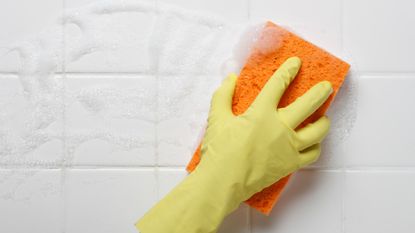 Person wearing a yellow rubber glove wiping down a bathroom wall