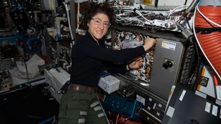 Christina Koch smiles at the camera while she works on hardware that consists of a lot of wires