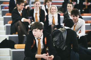 A scene set in the school where Kyle (Samuel Bottomley) is sitting at his desk at the front of the class looking over his shoulder nervously, while a group of girls behind him are looking at something on their phones and laughing