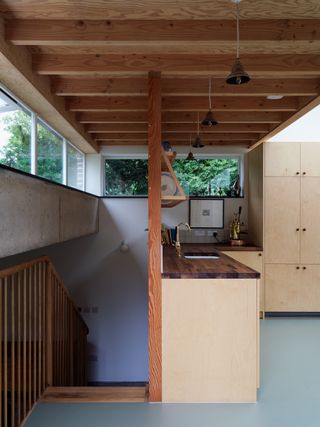 Interior of north London home extension showing two levels, bgrfey floor, wooden staircase and hand rail, white walls, kitchen area, wooden worktops, white sink and gold taps, light wood units, picture frame, windows with view of tree tops