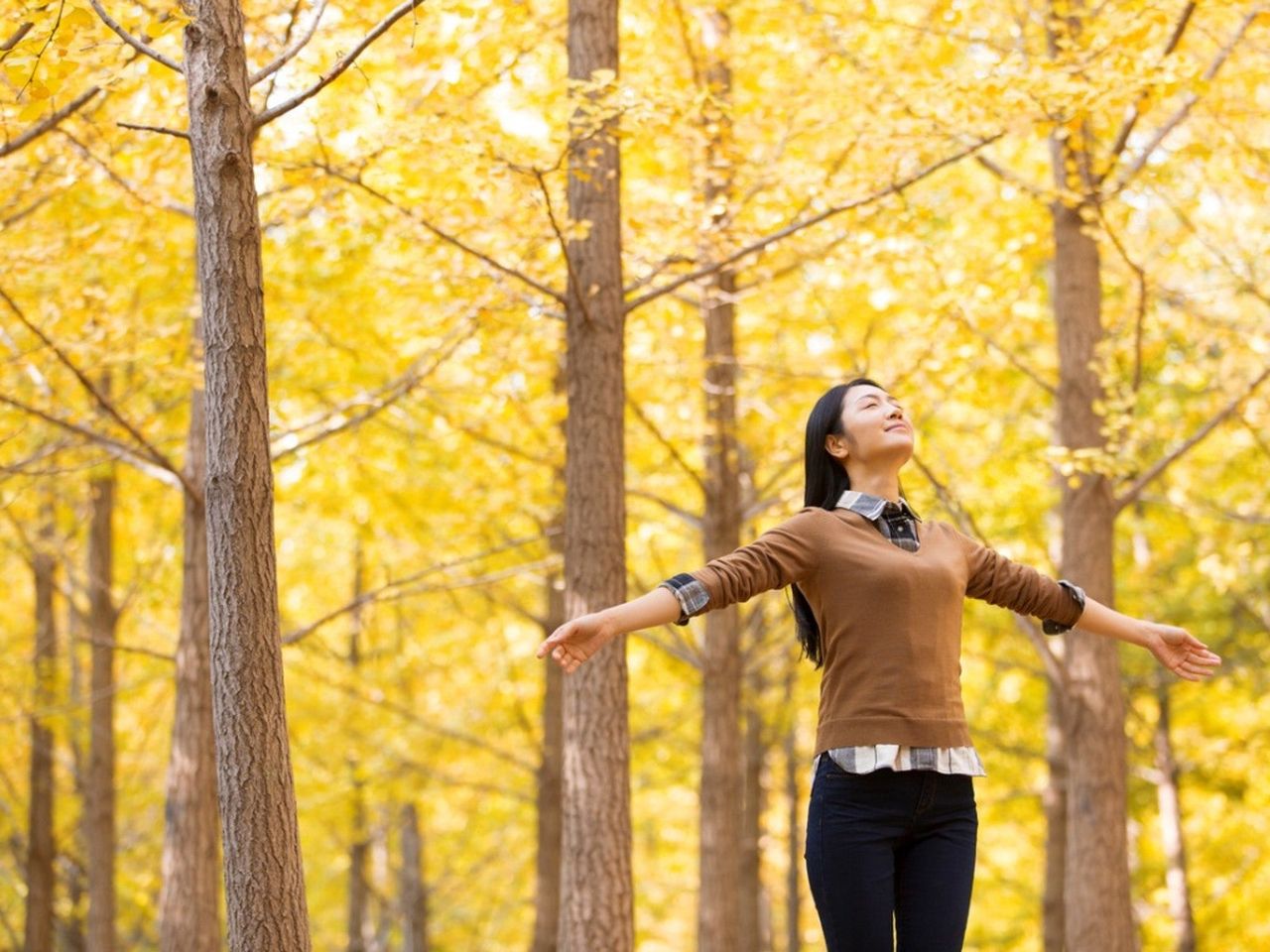 A woman stands with her eyes closed and arms outstretched in a forest of trees with yellow leaves