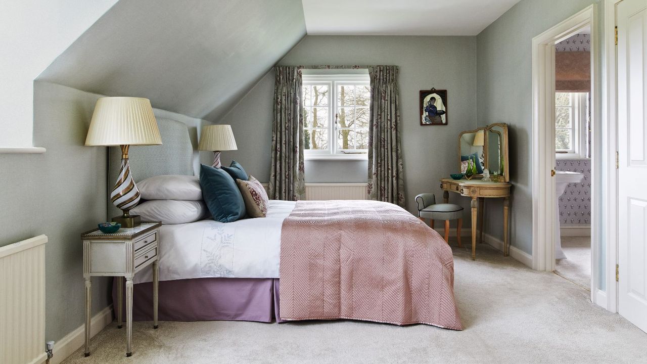 Peaceful bedroom with sloped ceiling, plush cream carpets and ornate gold dressing table with three-part mirror. There is a door open and a sliver of an en suite bathroom visible