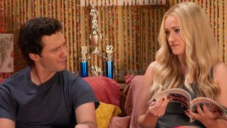 Georgie and Mandy discuss something in bed. She's reading a magazine and he has a confused look on his face. Both are fully clothed. 