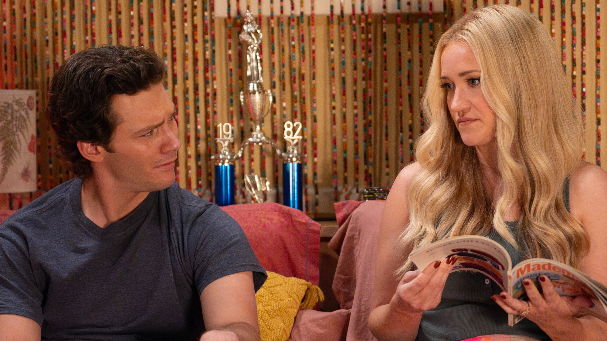 Georgie and Mandy discuss something in bed. She's reading a magazine and he has a confused look on his face. Both are fully clothed. 