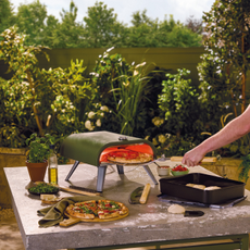 Aldi green pizza oven on stone table. A hand is putting a pizza into the oven, and there is a freshly cooked one next to it.