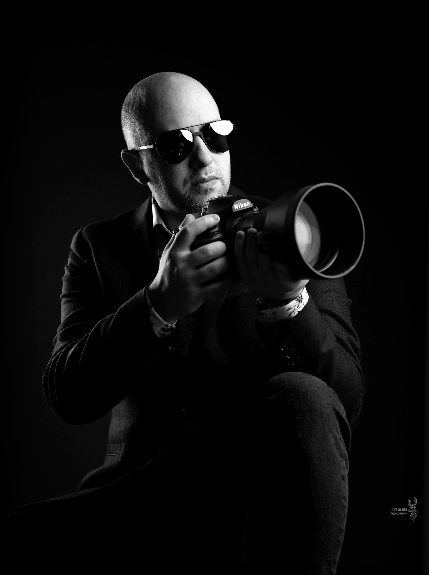Jon Segui holding a professional Nikon Z9 camera, dressed in a dark suit, against a black backdrop, exuding a serious, artistic vibe.
