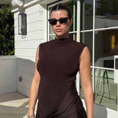 Sofia Richie Grainge wears a chocolate brown dress with a burgundy clutch bag and dark sunglasses.