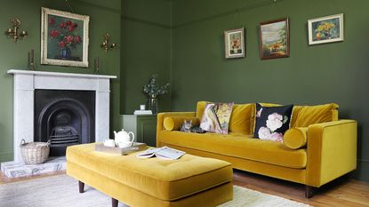 A green-painted living room with a yellow velvet sofa and matching ottoman and vintage framed paintings hanging on the walls