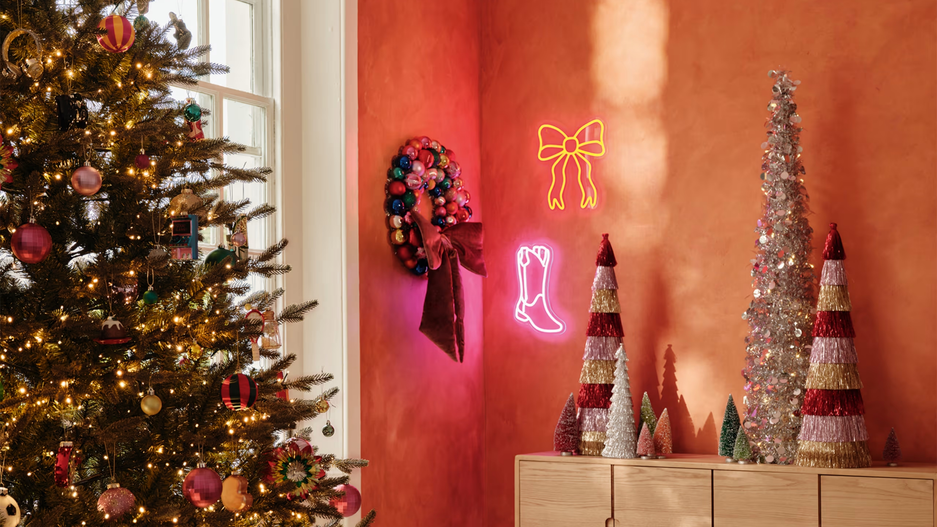 Image of an orange room with a christmas tree and a console table that has lots of miniature, sparkly Christmas trees on it. There are two neon lights hanging on the wall and a Christmas wreath.