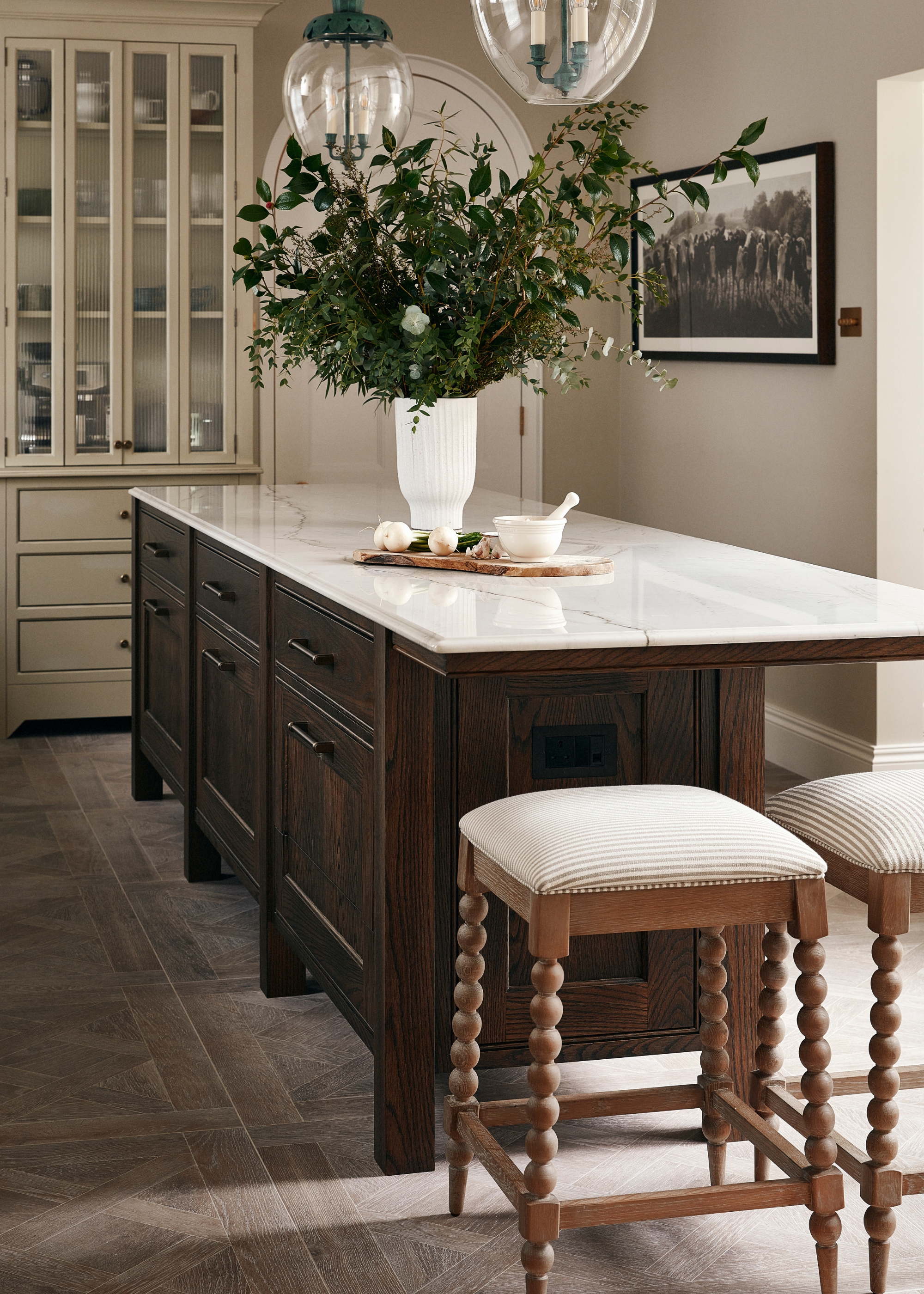 A modern traditional kitchen with a dark wood island in an off-white room, upholstered wooden stools and a big vase if greenery on the island.