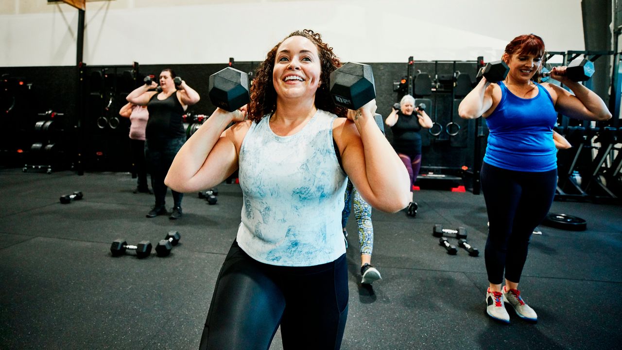 Smiling woman in gym class holding dumbbells by her shoulders. There are women in the background in the same position