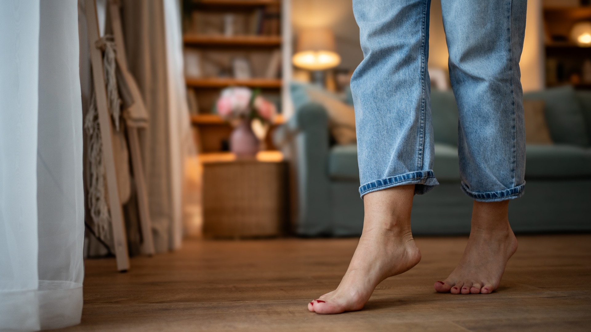 shot of bare feet in jeans on wooden floor with a living room scene blurred in the background. the feet are lightly on toes with red nails