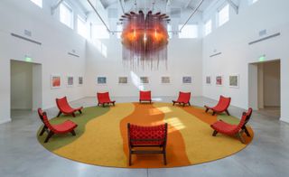 Exhibition view of Jorge Pardo ’Mongrel’. A room with red chairs around the edges of a round colourful rug, wall paintings and a large chandelier.