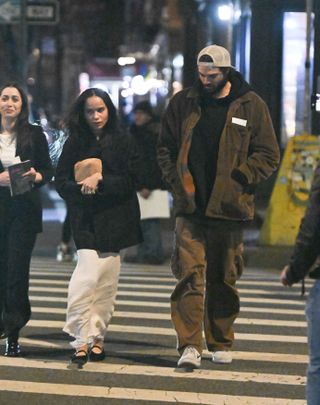 A photo of Zoë Kravitz accessorizing a white slip skirt with a black jacket, suede Mary Jane flats, and a textured brown clutch from The Row.