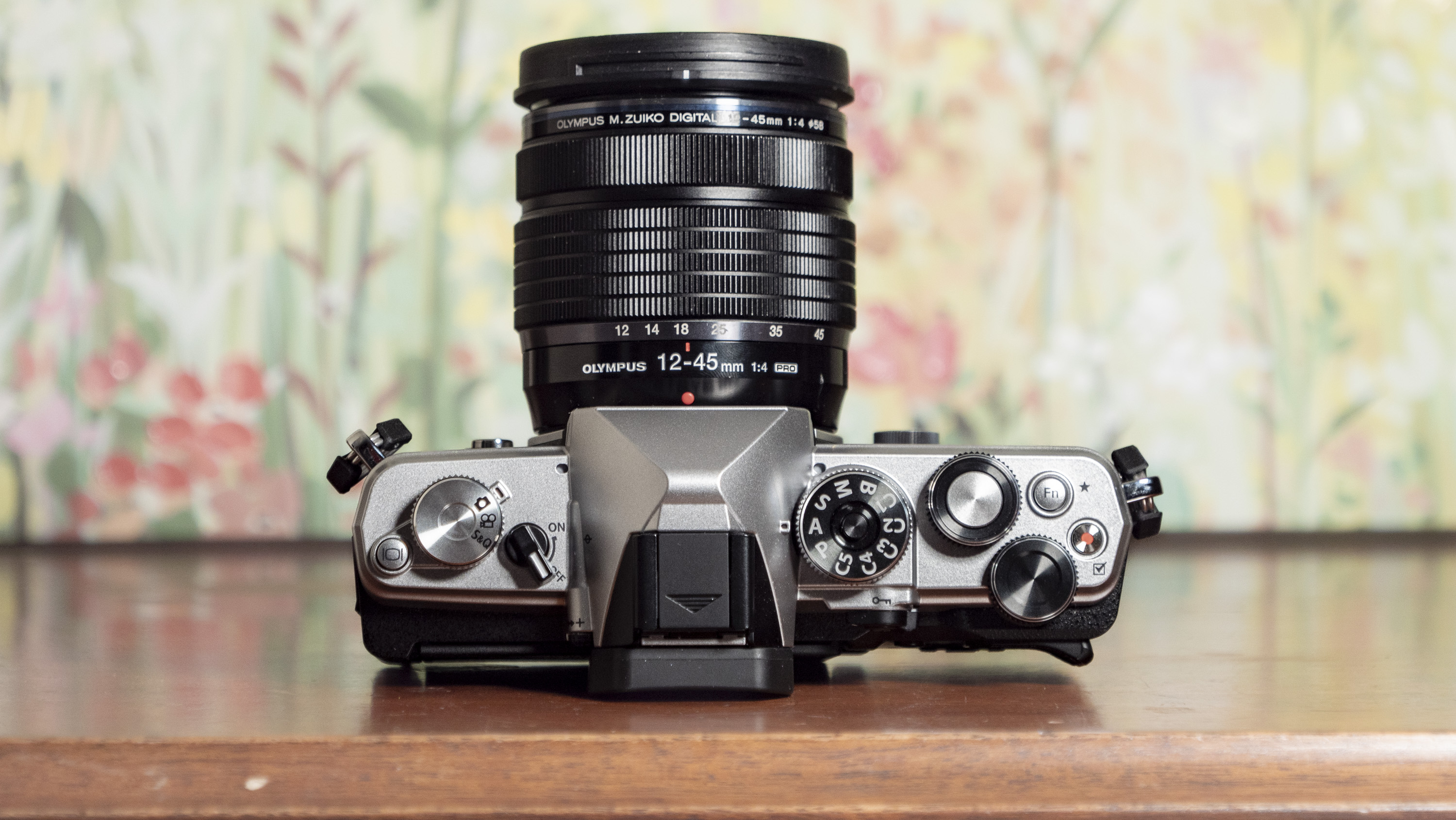 Top plate of the OM System OM-3 mirrorless camera on a wooden surface with floral painting background, 12-45mm F4 Pro lens attached