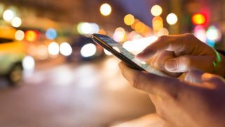 Hands in close up operate a cell phone while the lights of a city street at dusk glow in the background.