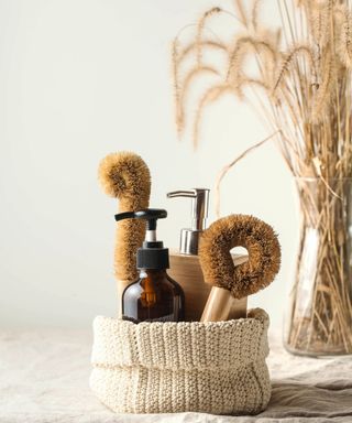 A knitted basket with glass dispensers and natural cleaning supplies inside, with dried flowers in the background