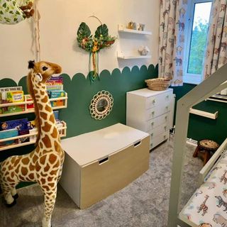 child's bedroom in a jungle theme with the bottom half of the walls in green with a curved pattern where it meets the top cream half, bookshelf on the wall behind a large toy giraffe, with a storage box and chest of drawers against the wall