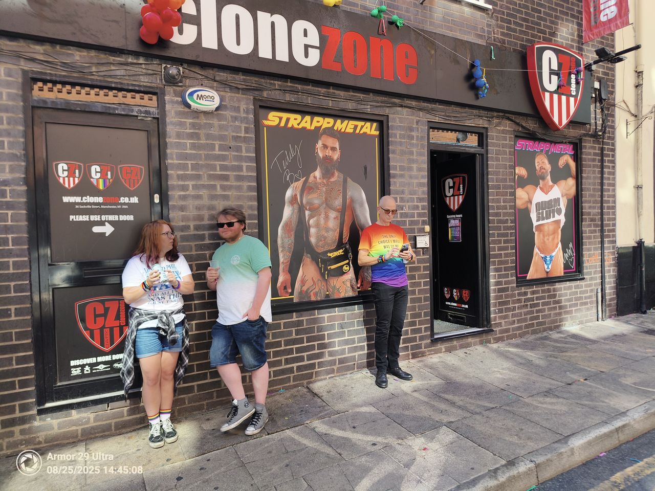 A group of three people stands on a sidewalk in front of a building with signs for "Clone Zone" and posters of muscular men.