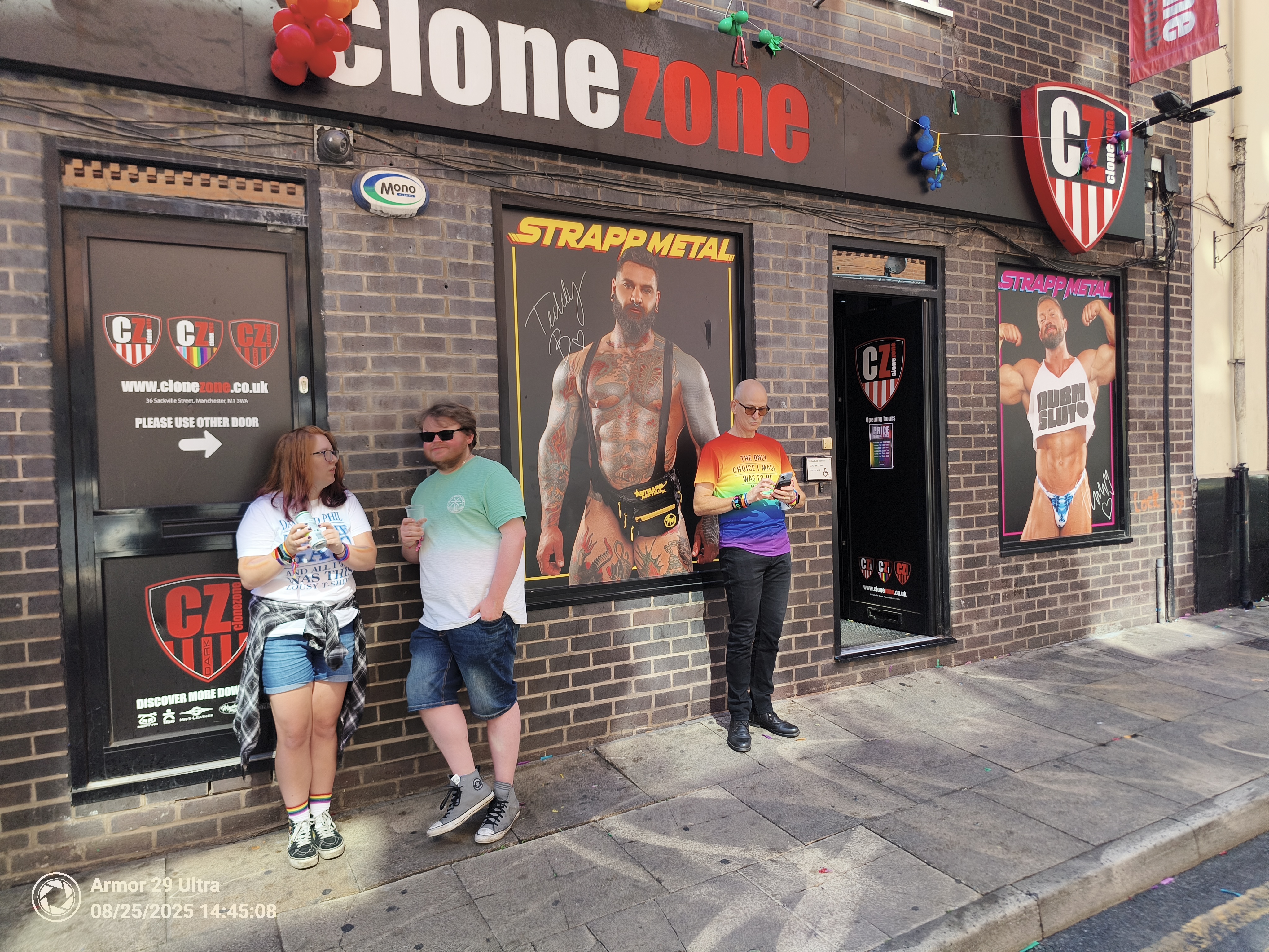 A group of three people stands on a sidewalk in front of a building with signs for "Clone Zone" and posters of muscular men.