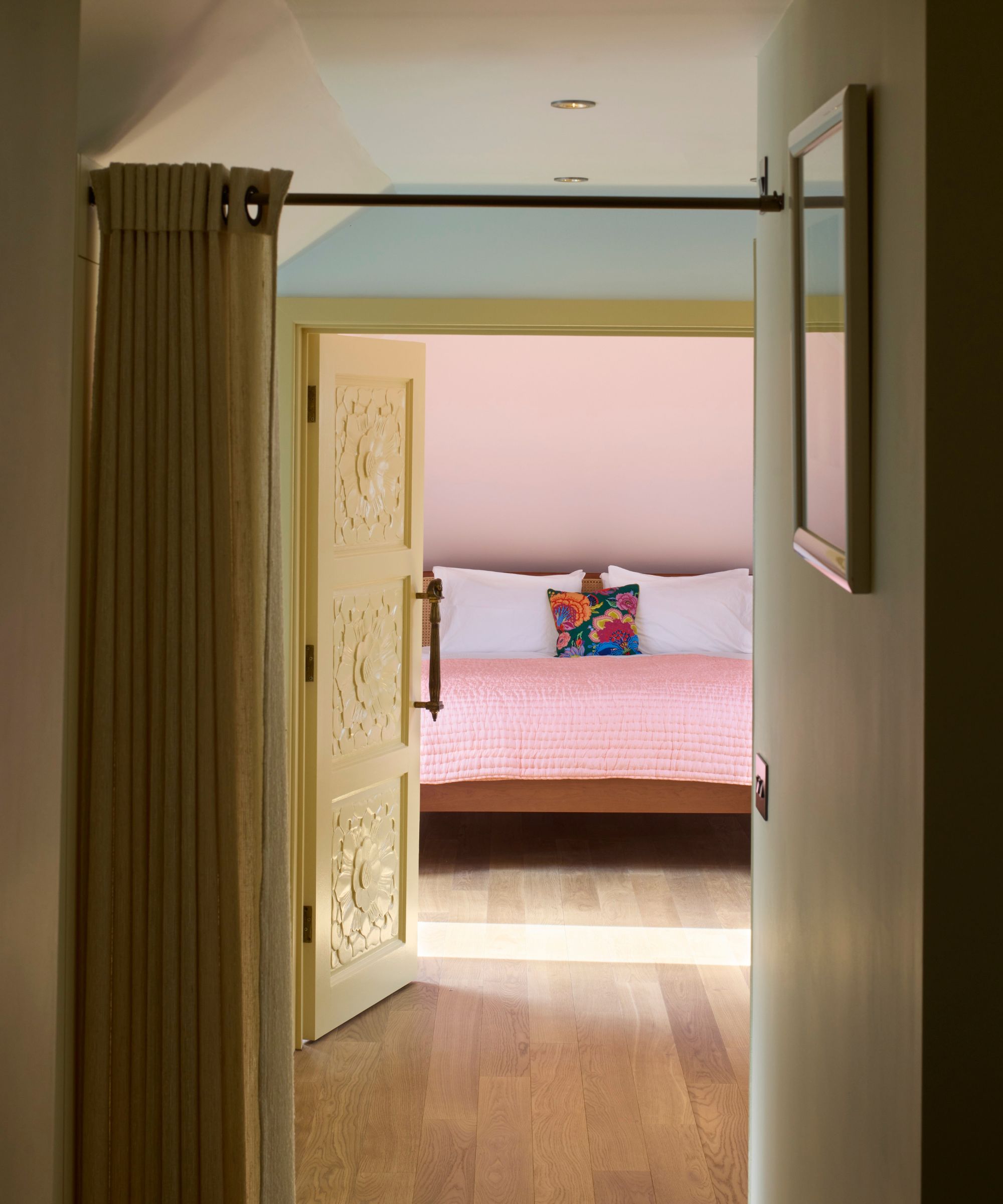 View through a doorway into a soft pink bedroom. A carved cream door stands open, revealing a bed dressed in a blush quilt with white pillows and a bright floral cushion. Light wood flooring runs from hallway into the sunlit room.