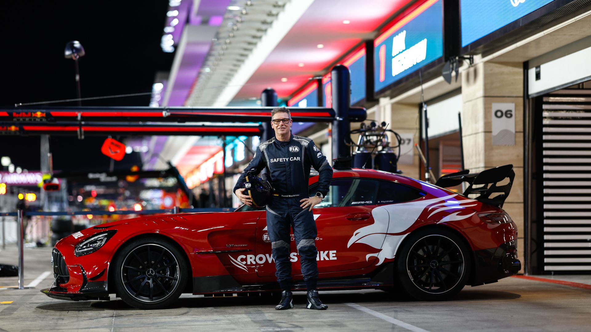 an image of Bernd standing by a red car in his driving gear, holding a racing helmet under his arm
