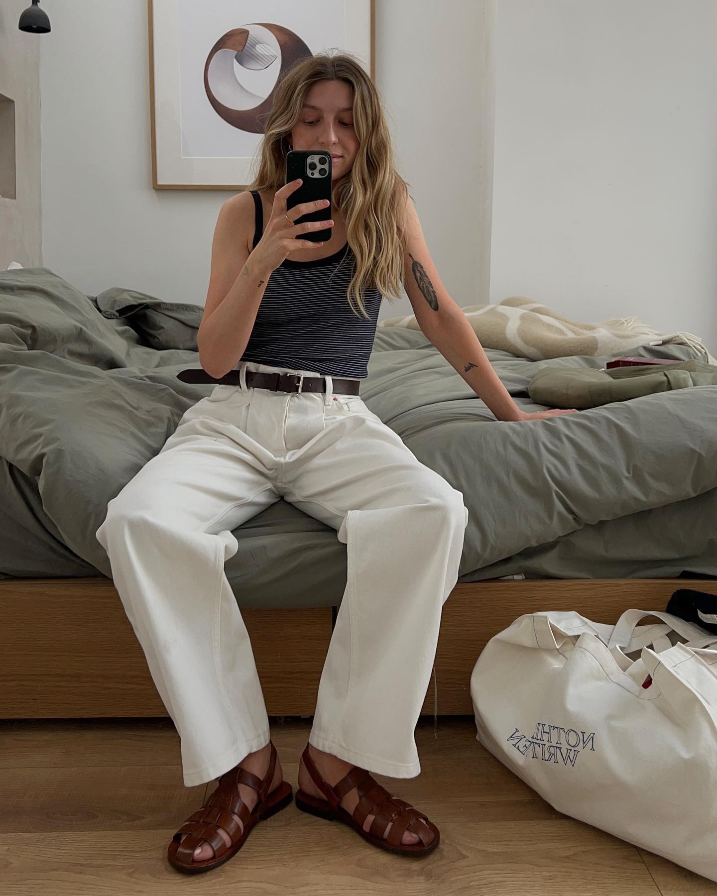 Influencer @brittanybathgate sits on a bed wearing white jeans with brown fisherman sandals and a black tank top.