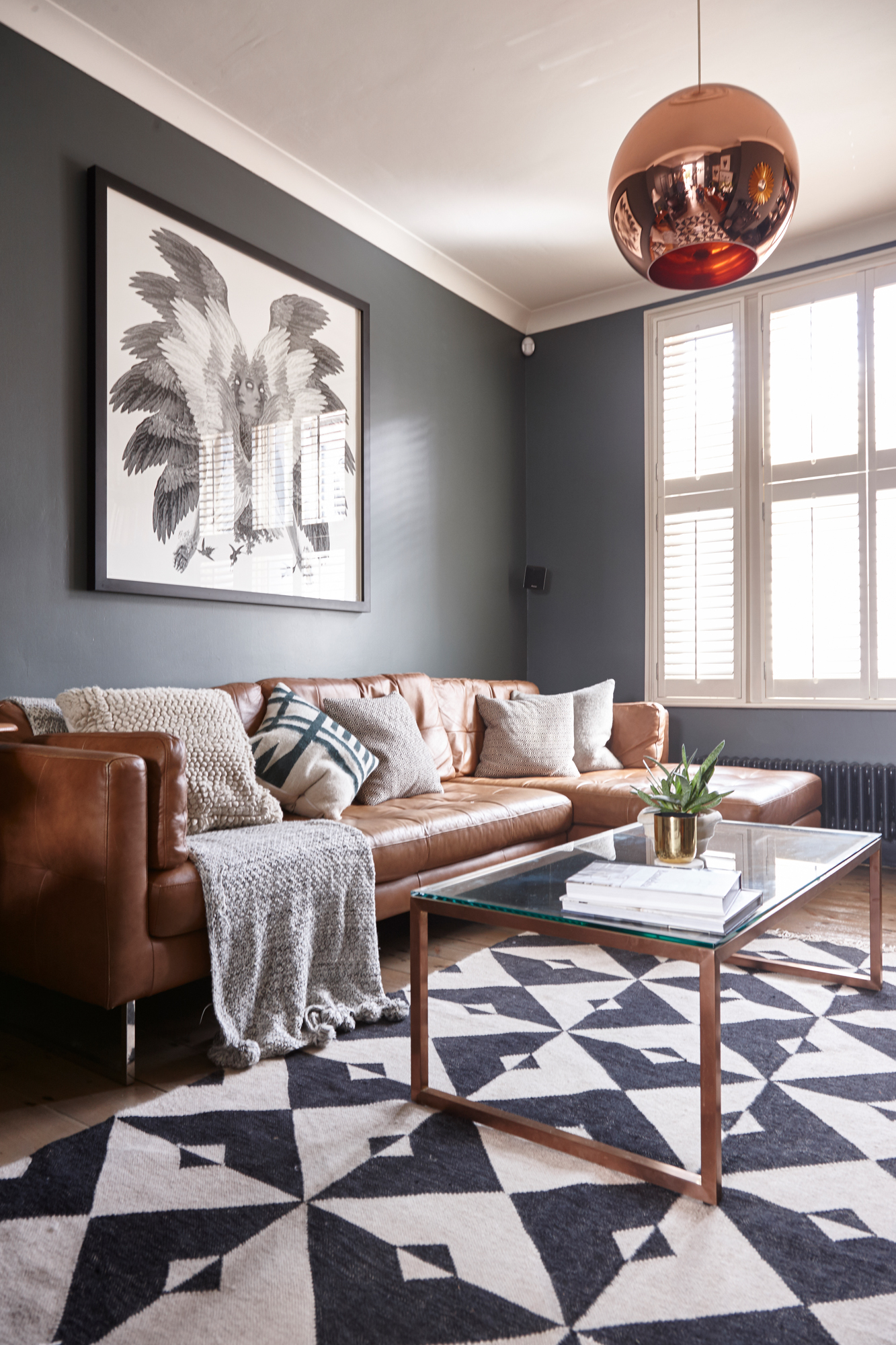 Jason Traves house: Living room with dark grey walls, tan leather sofa, minimalist coffee table with glass top, copper pendant light and black and white pattern rug over a wooden floor